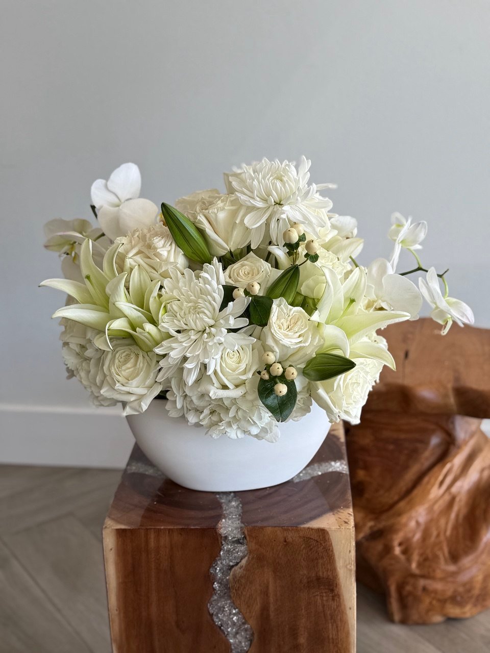 The arrangement is in a white, modern, bowl-shaped vase. It features a variety of white and cream-colored flowers, creating a monochromatic, elegant look.
